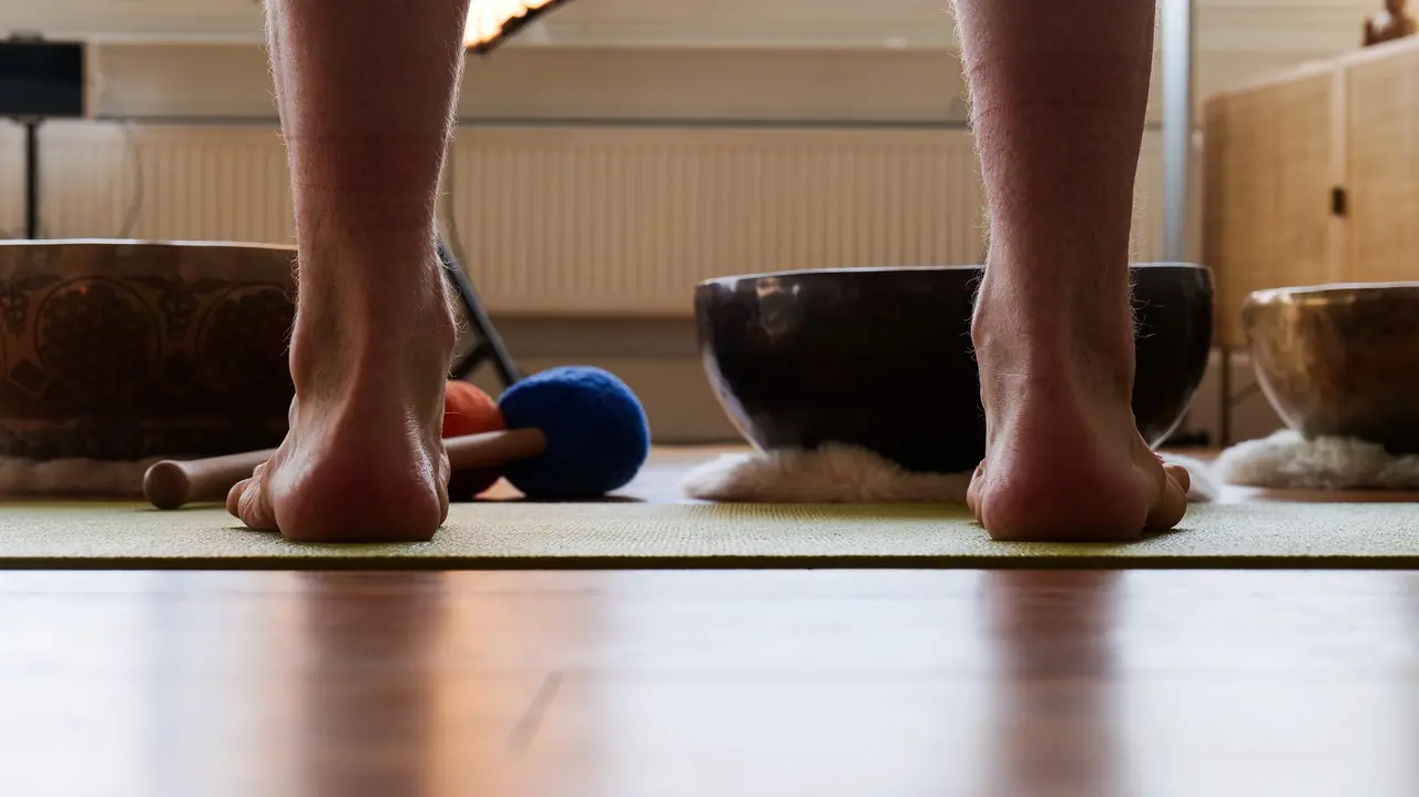 Zwei nackte Füße stehen auf einer Yogamatte, daneben Klangschalen auf dem Holzboden.