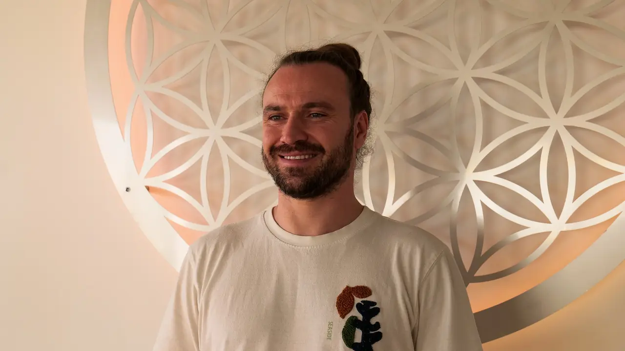 Michi Willkommen lächelt in die Kamera, hinter ihm die Blume des Lebens an der Studiowand.